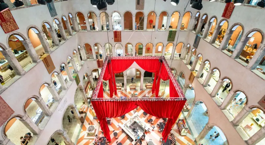 Interni del Fondaco dei Tedeschi con un'installazione di tende rosse al centro, che incornicia un'area bar e tavoli. Le arcate dei piani superiori ospitano negozi.