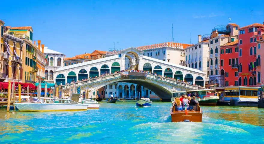 Gondole e vaporetti solcano il Canal Grande, sotto il Ponte di Rialto affollato di gente. Un assaggio delle **esperienze Venezia**.