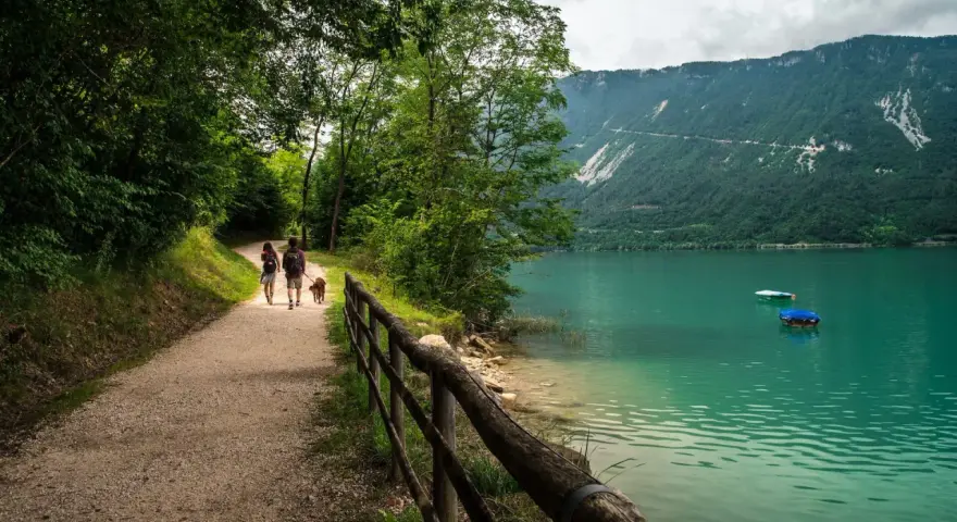Coppia con cane passeggia lungo il sentiero del lago di Santa Croce, godendosi la natura e il panorama montano.