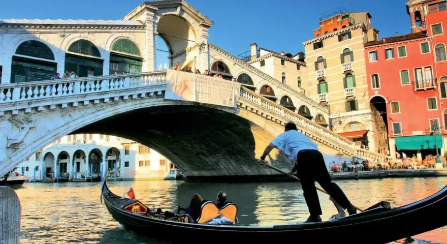 Gondola sotto il Ponte di Rialto, un'icona delle piazze Venezia, con edifici colorati e turisti che ammirano il panorama.