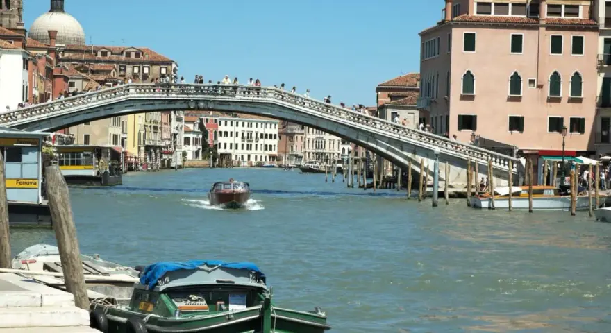 Un motoscafo naviga sotto uno dei ponti di Venezia, con edifici storici e gente che passeggia.