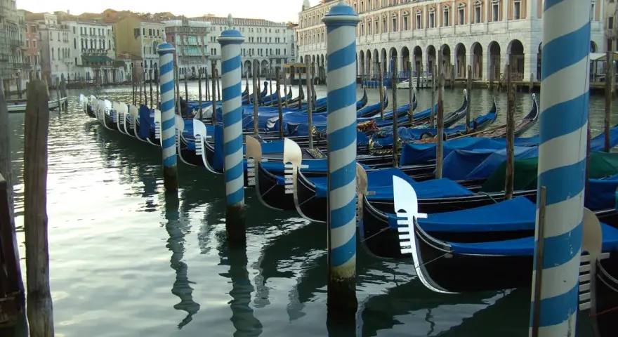 Una fila di imbarcazioni tipiche veneziane, le gondole, ormeggiate lungo un canale, con i loro caratteristici pali a strisce blu e bianche.