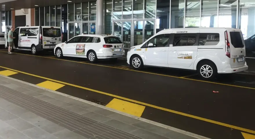 Taxi a Venezia, in attesa all'aeroporto. Diverse auto bianche, tra cui minivan e station wagon, pronte per il trasporto passeggeri.