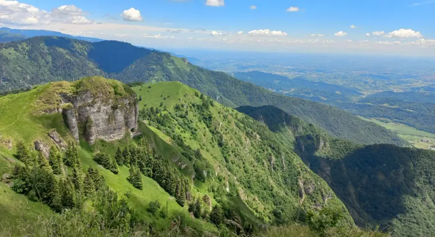 Panoramica mozzafiato dal Passo San Boldo, con verdi pendii montuosi che scendono verso una valle lontana sotto un cielo azzurro con nuvole bianche.