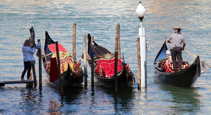 Gondole veneziane ornate, una turista scatta foto. La differenza tra gondola e sandalo è chiara: una naviga, l'altro si indossa.