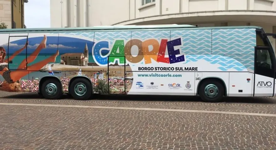 Bus turistico con grafica promozionale di Caorle, borgo storico sul mare. Immagini di spiaggia, mare e una donna in bikini.