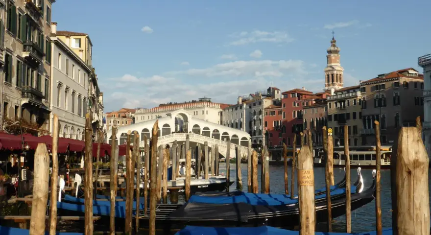 Gondole ormeggiate nel Canal Grande, con il Ponte di Rialto sullo sfondo. Un'immagine di Venezia romantica, con palazzi storici e il cielo azzurro.