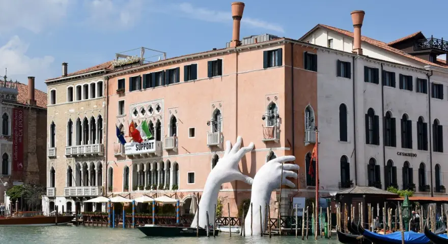 Enorme installazione di mani bianche emerge dal Canal Grande, davanti al museo arte contemporanea Venezia. Gondole ormeggiate.