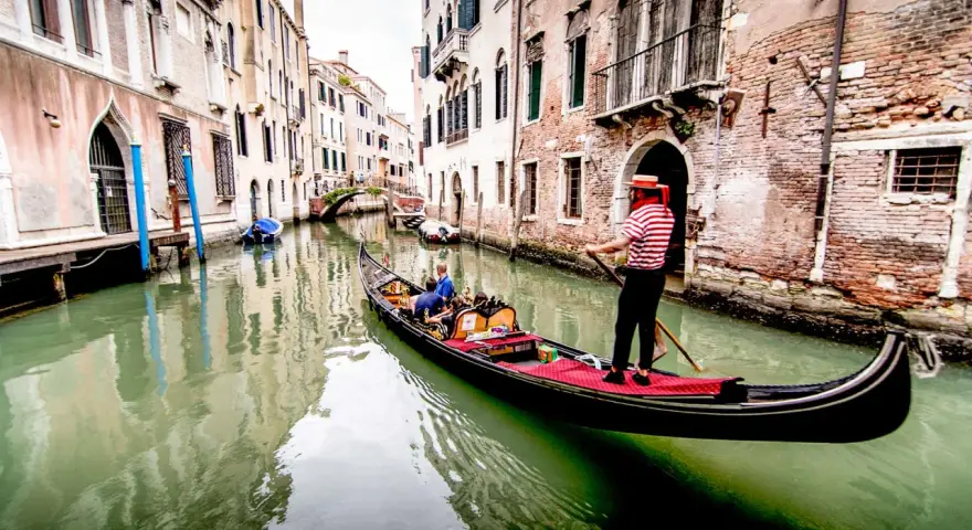 Gondoliere in abito a righe guida una gondola con turisti lungo un canale veneziano. Chissà quanto costa un giro in gondola a Venezia per vivere questa magia.
