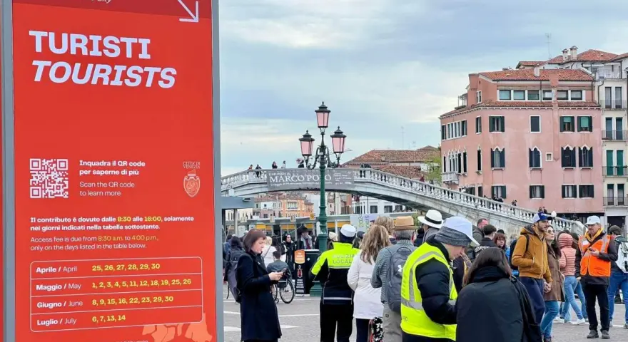 Cartello rosso a Venezia per il contributo accesso turisti, con date e QR code. Persone in fila sullo sfondo.