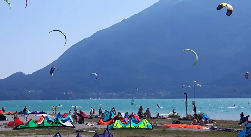 Coloratissima spiaggia sul lago di Santa Croce, con kitesurfisti che sfrecciano sull'acqua turchese e vele colorate che ondeggiano al vento.