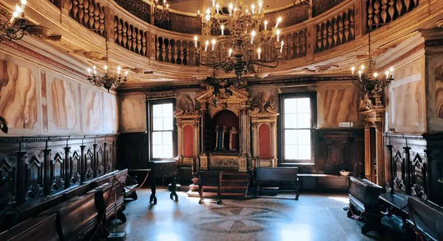 Interno sontuoso della sinagoga di Venezia, con arredi in legno scuro, un grande lampadario e l'Arca Santa dorata.
