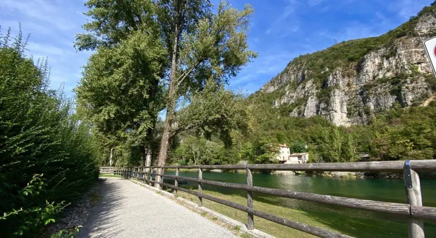 Sentiero ghiaioso lungo il fiume, con alberi verdi e una montagna rocciosa sullo sfondo. La **ciclabile del Brenta** invita a una passeggiata.