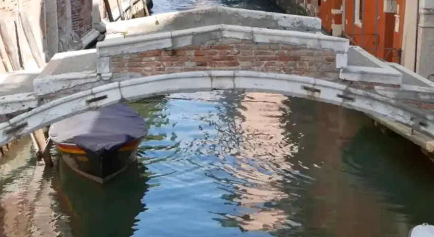Un piccolo ponte a Venezia, senza parapetto, sovrasta un canale dove una barca coperta attende.