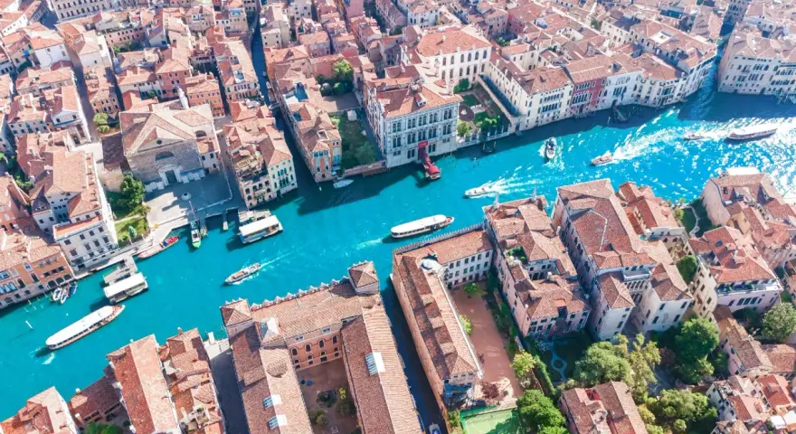 Vista aerea del **canale di Venezia** con gondole e vaporetti che navigano tra palazzi storici dai tetti rossi.