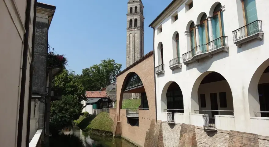 Canale e campanile a Oderzo, cosa vedere: un'elegante facciata bianca con portici e balconi si affaccia sull'acqua, con un ponte ad arco in mattoni.