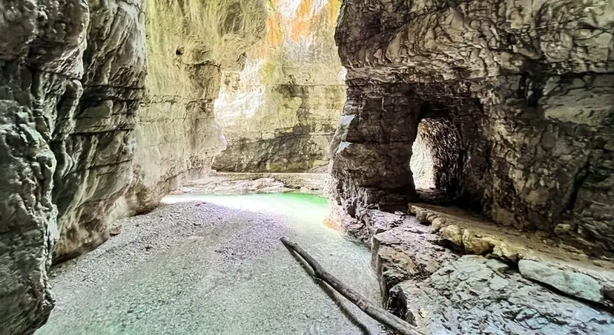 Gola rocciosa con acqua cristallina che ricorda il lago del Mis. Un sentiero scavato nella roccia conduce verso un'apertura luminosa.