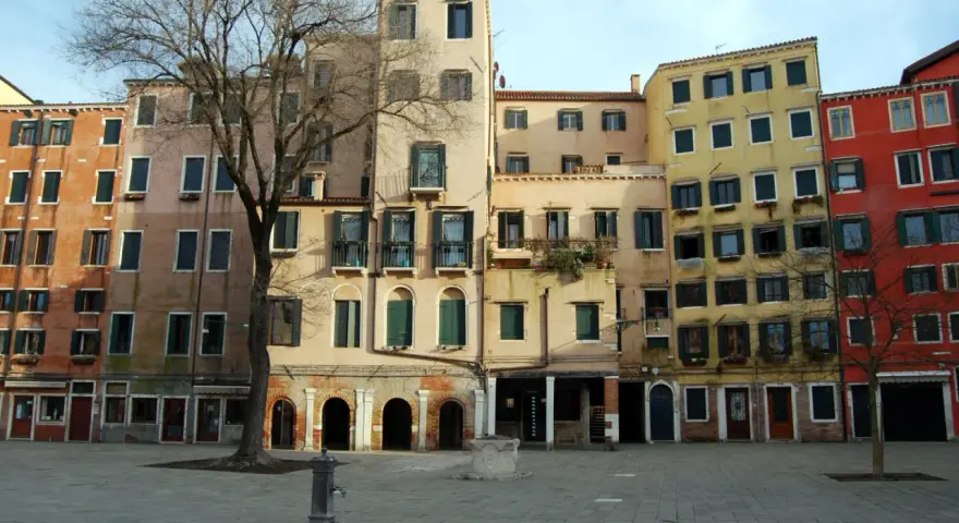 Edifici colorati nel ghetto ebraico di Venezia, con un albero spoglio e una fontana nella piazza lastricata.
