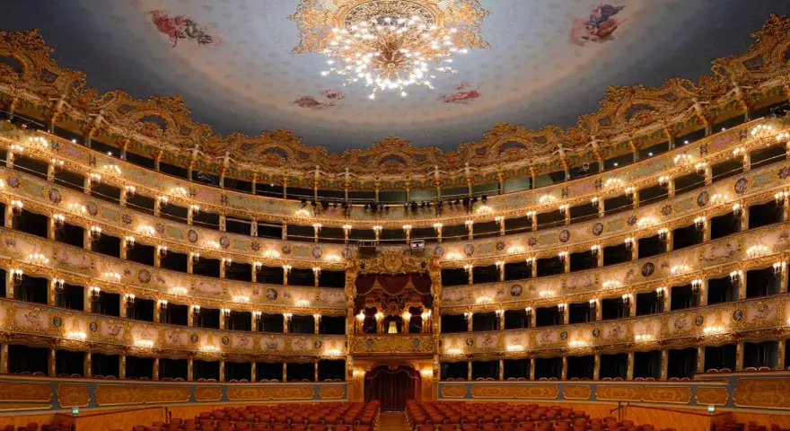 Interno sfarzoso del teatro La Fenice a Venezia, con palchi dorati, affreschi sul soffitto e un lampadario scintillante.