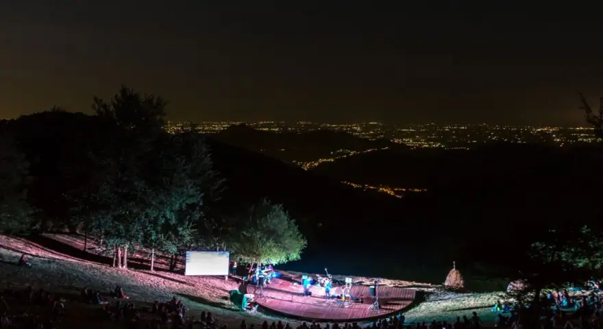 Concerto serale all'anfiteatro del Venda, con un pubblico seduto sull'erba e una band sul palco illuminato, sullo sfondo di un panorama notturno di luci cittadine.
