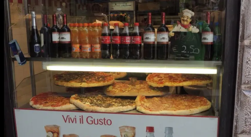 Vetrine con pizze, bottiglie di vino e bibite, un cartello "PIZZA DA € 2.50" e un cuoco decorativo. Sembra un ottimo posto per trovare le migliori pizzerie a Venezia.
