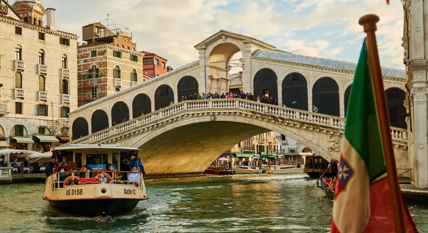 Traghetto della linea 2 di Venezia naviga sul Canal Grande sotto il Ponte di Rialto, con gondole e edifici storici sullo sfondo.