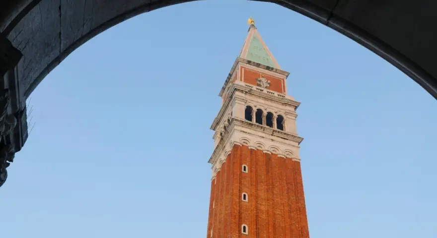 Vista dal campanile di San Marco: la torre in mattoni rossi si staglia contro il cielo azzurro, incorniciata da un arco di pietra.