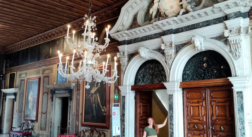 Una donna in un abito verde posa davanti a un portale decorato in una casa museo a Venezia, con un lampadario di cristallo e dipinti alle pareti.