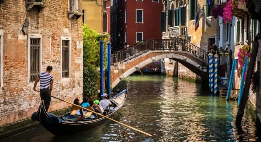 Gondola con turisti naviga in un canale, sotto un ponte, scoprendo la Venezia segreta tra palazzi antichi e fiori.