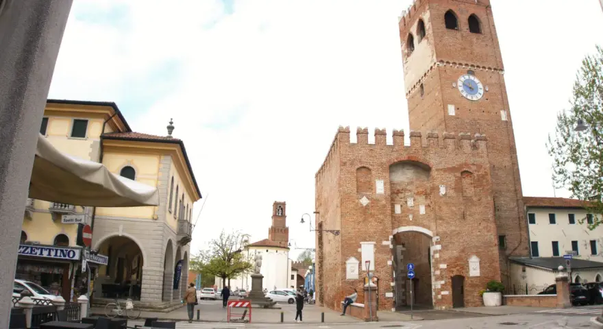 Piazza con torre medievale, portici e negozi. Ecco cosa vedere a Noale: la torre civica con orologio, il palazzo giallo e i tavolini all'aperto.