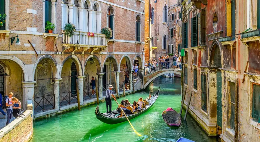 Un gondoliere a Venezia guida una gondola con turisti lungo un canale verde, tra palazzi storici e un ponte affollato.