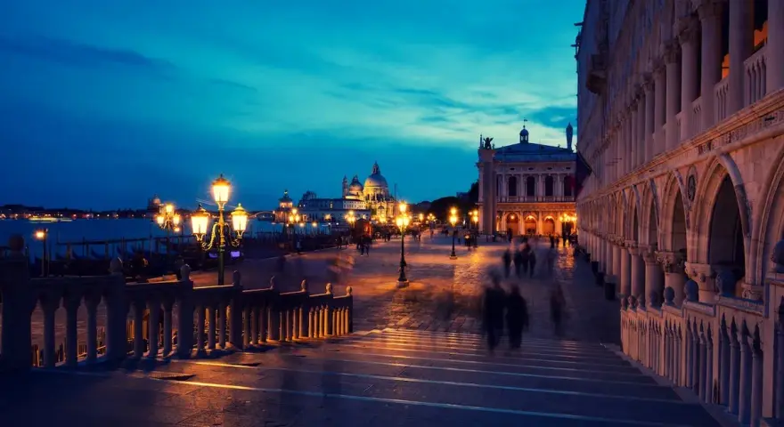 Venezia di notte, con le luci dei lampioni che illuminano la piazza e le sagome sfocate dei passanti.