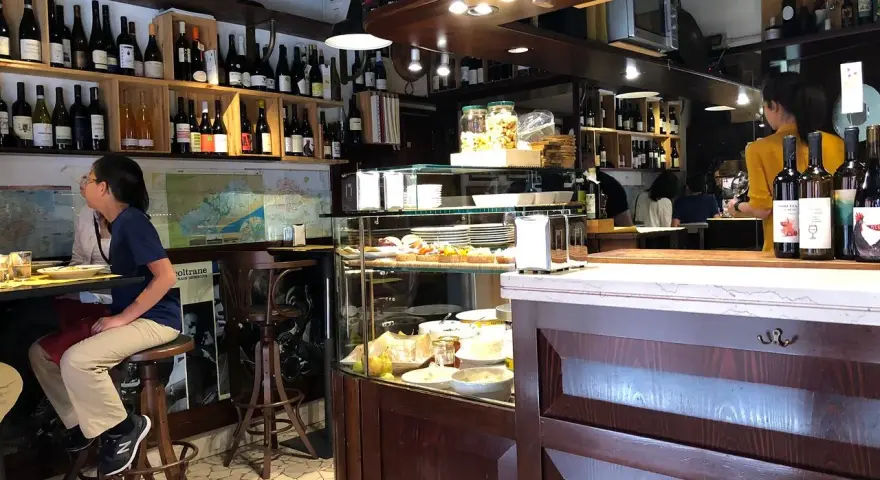 Interno di un'osteria al cicheto a Venezia, con bottiglie di vino, cibi in vetrina e persone sedute al bancone.