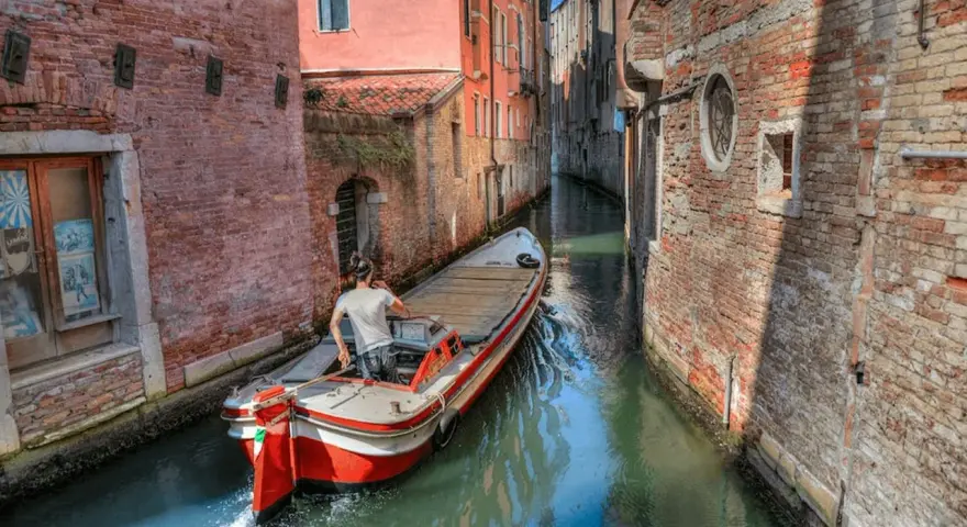 Un uomo guida una barca su un canale stretto a Venezia, circondato da edifici in mattoni. Quanti canali ci sono a Venezia? Molti, come questo!