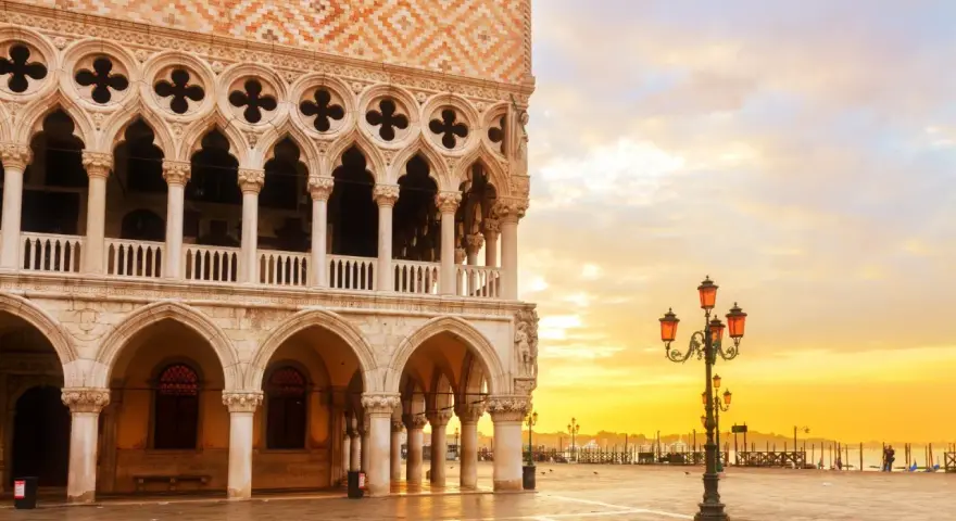 Palazzi storici di Venezia all'alba, con il sole che illumina la piazza e le architetture gotiche.