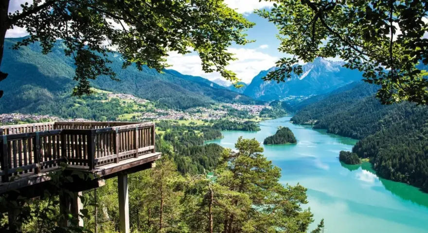 Terrazza panoramica sui laghi del Cadore, con montagne sullo sfondo e un'isola verde nel lago turchese.