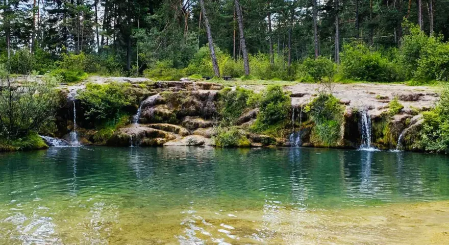 Cascate naturali nella valle di Cadore, con acqua cristallina che scende tra rocce e vegetazione rigogliosa, circondate da un bosco di pini.