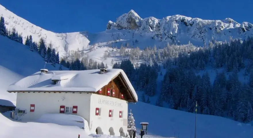 Rifugio innevato nelle Dolomiti, ideale per ciaspolate invernali. Montagne maestose e boschi coperti di neve.