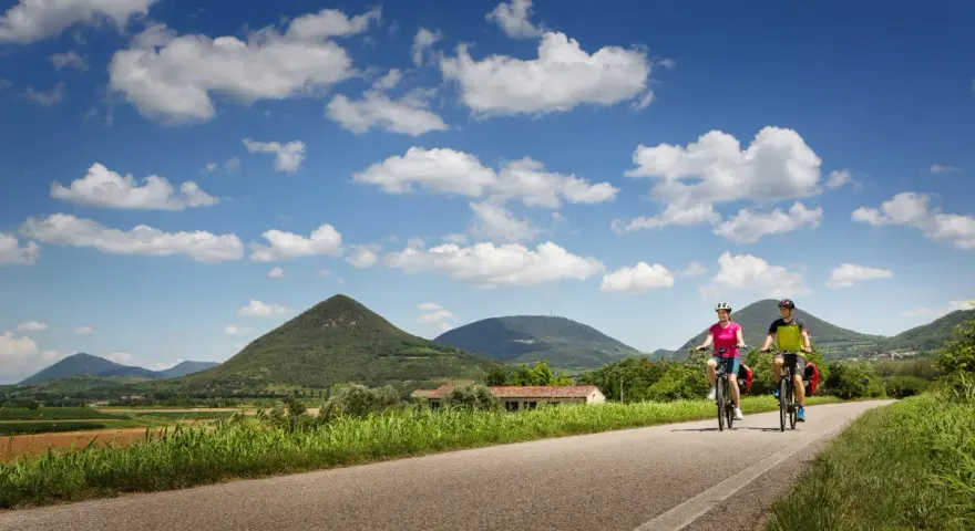 Coppia in bicicletta lungo la ciclabile dei Colli Euganei, tra verdi colline e cielo azzurro con nuvole bianche.