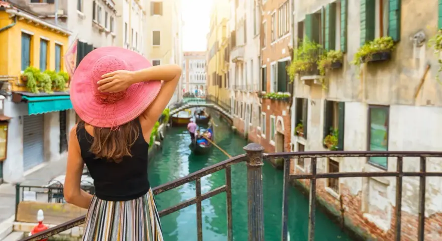 Donna con cappello rosa ammira un canale a Venezia, con gondole e palazzi storici. Un'esperienza da vivere a piedi, tra Santa Lucia e Piazza San Marco.