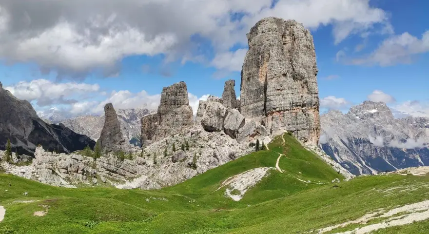 Sentiero che serpeggia tra verdi prati verso le maestose Cinque Torri Dolomiti, sotto un cielo azzurro con nuvole bianche.