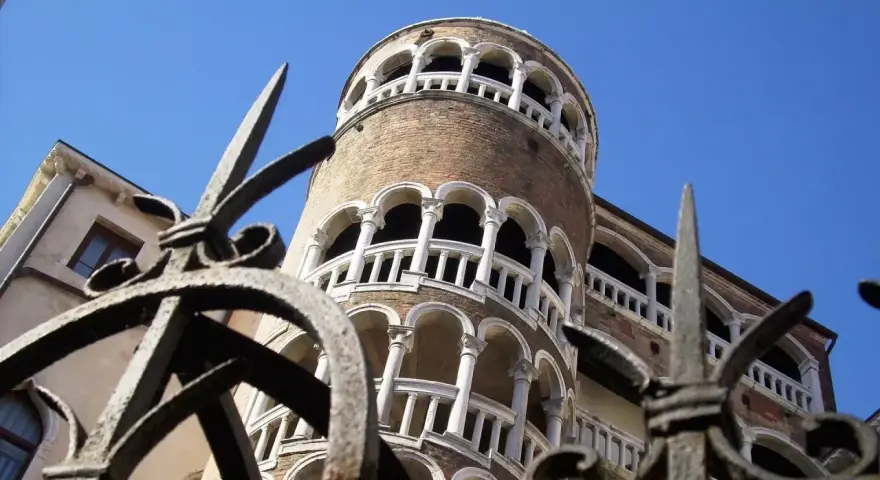La Torre del Bovolo, con la sua architettura a chiocciola e i suoi loggiati, si staglia contro il cielo azzurro, vista attraverso un cancello in ferro battuto.