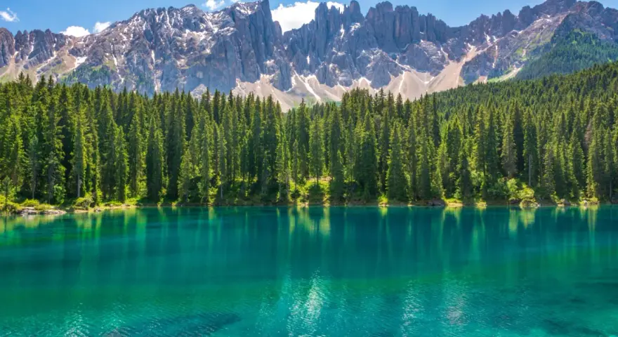 Lago alpino dalle acque turchesi, circondato da una fitta foresta di abeti, con le maestose cime delle Dolomiti innevate sullo sfondo. Un paesaggio da sogno, tra Venezia e le Dolomiti.