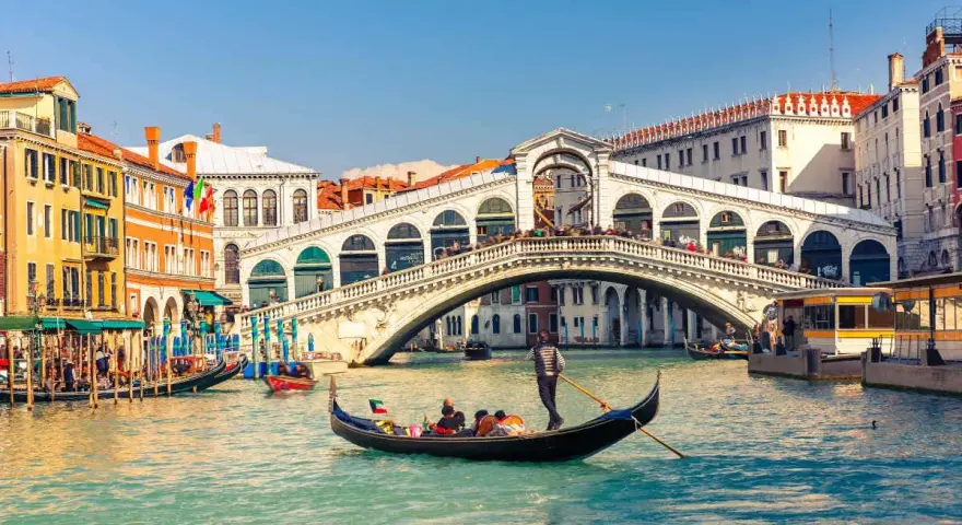 Una gondola solca il Canal Grande a Venezia, passando sotto il Ponte di Rialto, con edifici storici e altre imbarcazioni sullo sfondo.