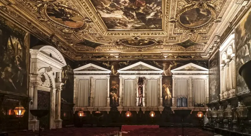 Sala capitolare Scuola Grande di San Rocco: soffitto dorato affrescato, pareti con dipinti e statue, sedie rosse disposte per un evento.