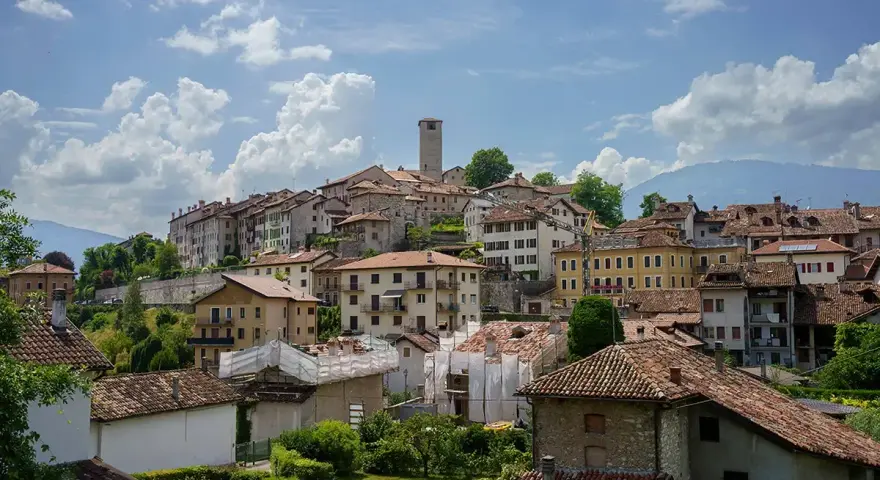 Panoramica di Feltre e dintorni, con case dai tetti rossi che si arrampicano sulla collina sotto un cielo azzurro e nuvoloso.
