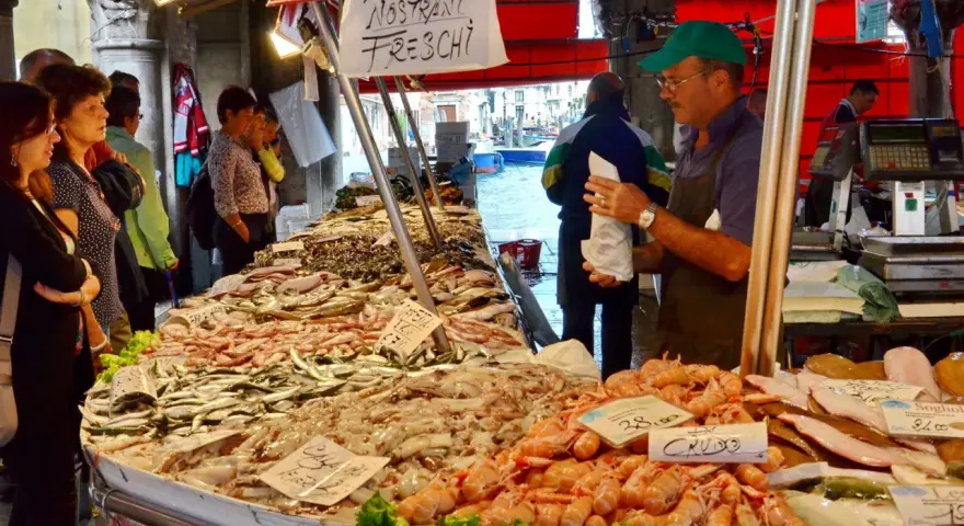 Mercato Venezia: bancarella di pesce fresco con gamberi, calamari e pesciolini. Venditore con cappellino verde.
