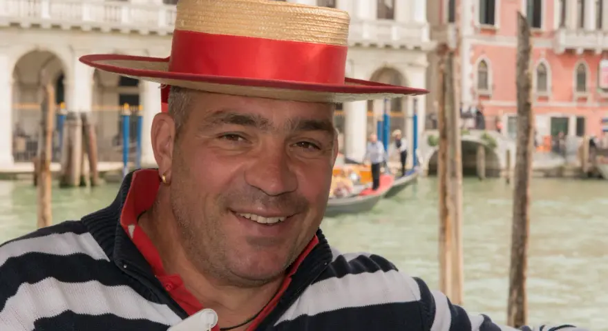 Un gondoliere sorridente, con il suo cappello iconico, pronto a navigare i canali di Venezia, la città che porta il soprannome di "La Serenissima".