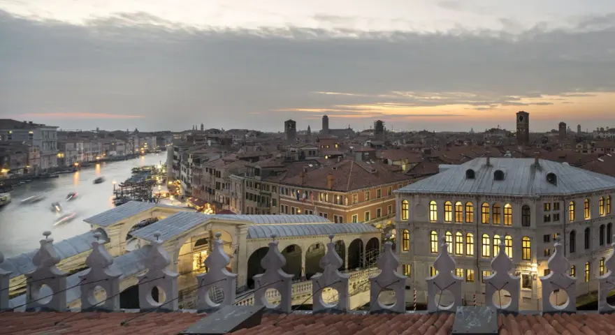 Vista dal **fondaco dei tedeschi Venezia terrazza** al tramonto, con il Canal Grande e il Ponte di Rialto illuminati.