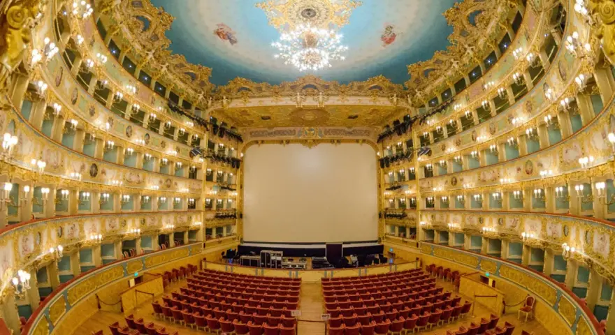 Interno sfarzoso del Teatro La Fenice di Venezia, con palchi dorati, soffitto affrescato e un grande schermo bianco.